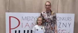 Klara Utrata stoi ze swoją nauczycielką Jagodą Młynarską na tle planszy konkurs pianistyczny za kotarą
