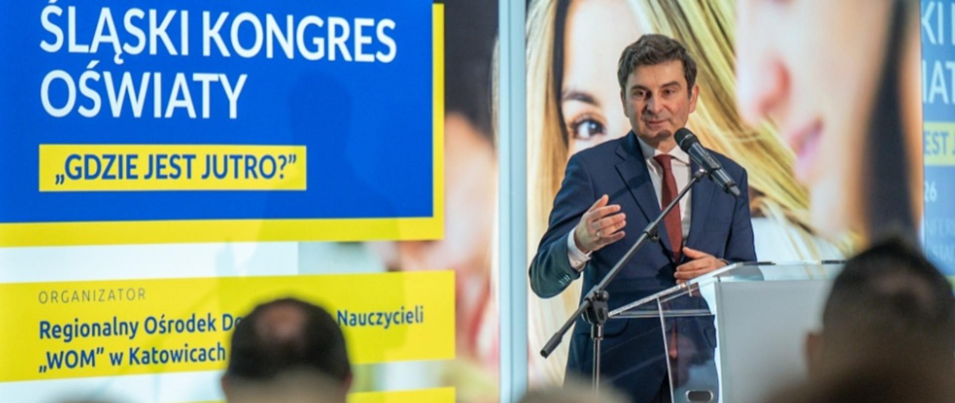 Mężczyzna w garniturze przemawia przy mównicy z mikrofonem podczas konferencji. Za nim widoczny duży baner z napisem „Śląski Kongres Oświaty – Gdzie jest jutro?” oraz osoby siedzące na widowni.