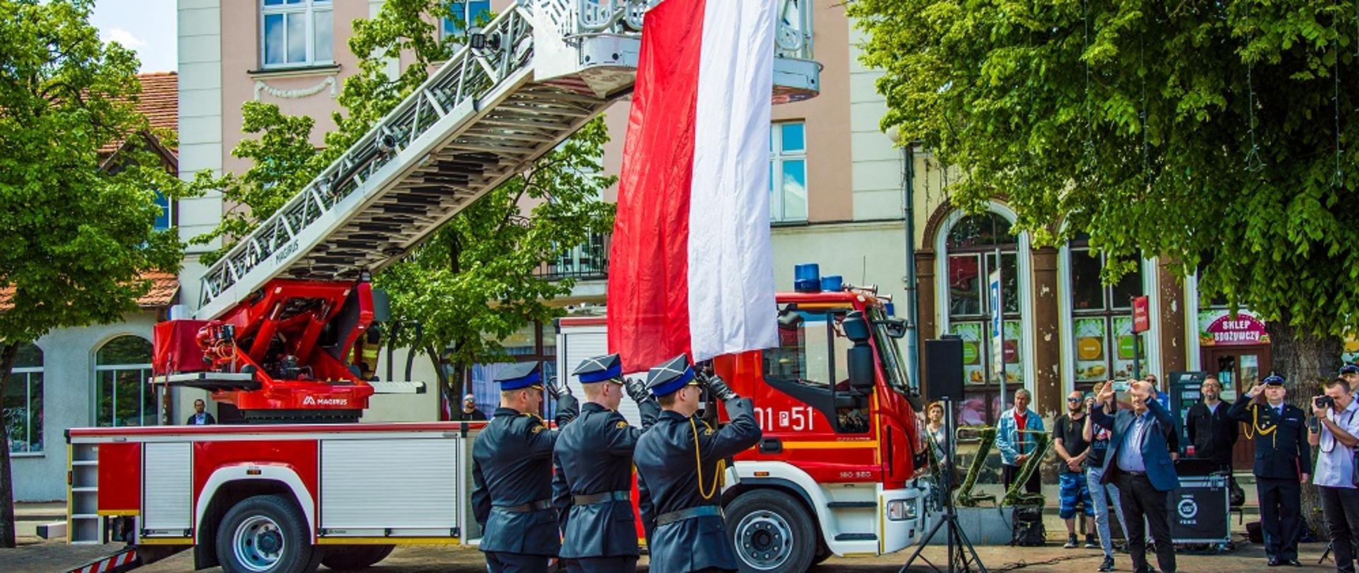 Zdjęcie przedstawia strażaków podczas święta strażaków powiatu chodzieskiego oraz miasta Chodzież.
W tle budynki i samochód strażacki.
