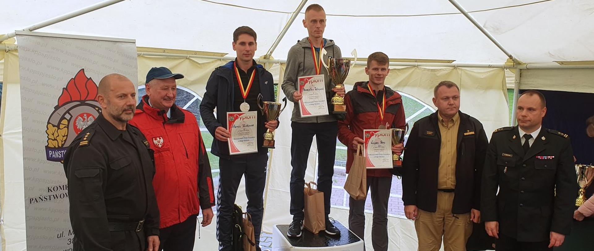 Na zdjęciu 3 osoby ubrane w stroje sportowe stojące na podium z medalami, dyplomami i pucharami. Obok nich strażacy w mundurach. W tle widać baner z napisem Komenda Powiatowa Państwowej Straży Pożarnej w Jaśle. 