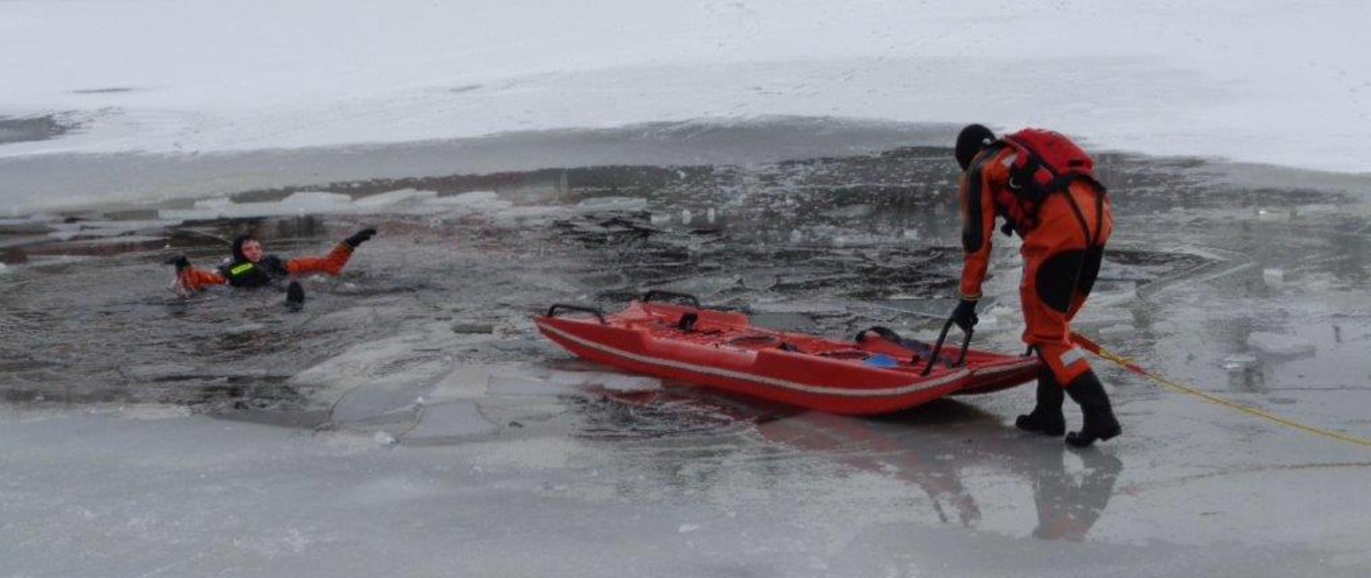Próba podjęcia osoby poszkodowanej na lodzie przy pomocy sań lodowych, strażaka w zabezpieczeniu.