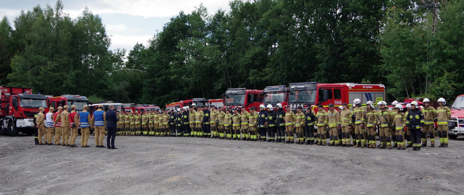 Widok z przodu. Zbiórka uczestników ćwiczeń. W tle pojazdy pożarnicze.