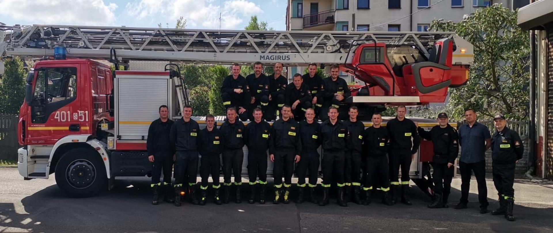 Grupa strażaków ubranych w mundury koszarowe na tle samochodu specjalnego pożarniczego - drabiny 