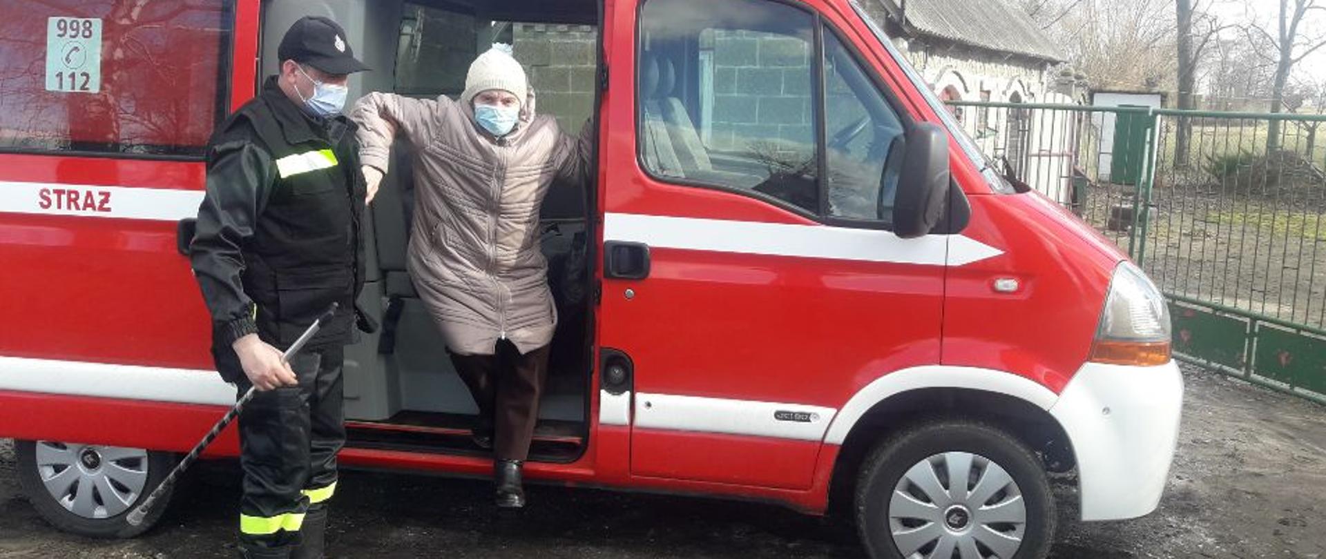 Strażak w ubraniu koszarowym lewą ręką pomaga wysiąść z samochodu pożarniczego osobie starszej, w prawej ręce trzyma laskę. 