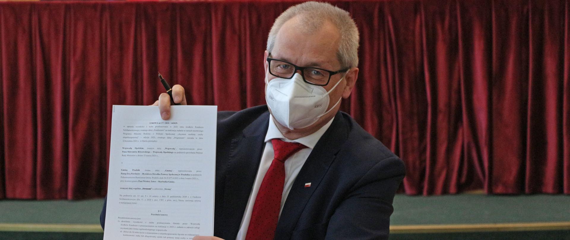 Na zdjęciu za stołem, siedzi wojewoda trzymający w rękach dokument. Pozuje do zdjęcia, prezentując dokument. Wojewoda spogląda w obiektyw, na twarzy ma maseczkę i okulary. W tle widać podest koloru zielonego i w oddali stół. 