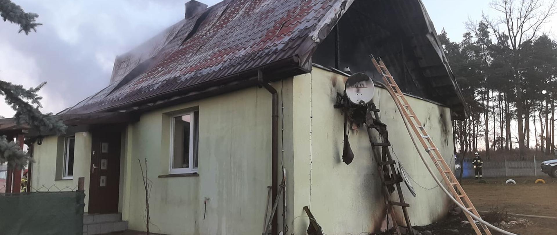 Zdjęcie przedstawia dom jednorodzinny, w którym spaleniu uległo poddasze. Brak ściany szczytowej. O budynek oparte są drabiny. Z dachu wydobywa się dym. 