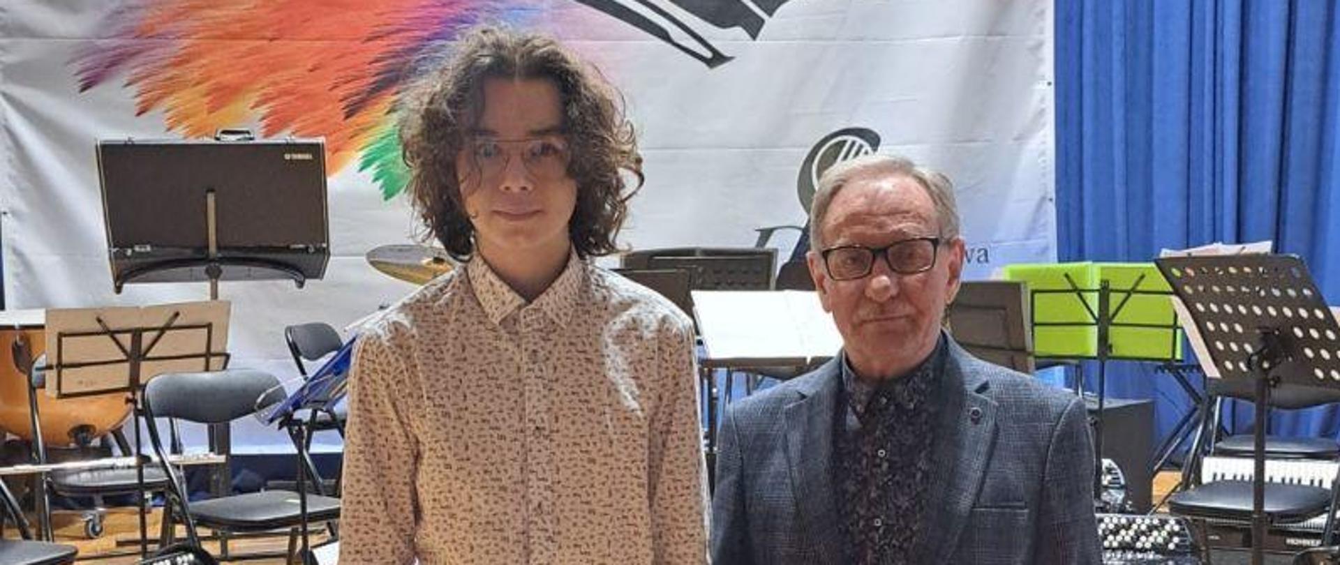 Zdjęcie kolorowe. Dwie osoby w strojach galowych na scenie. Od lewej stoją: uczeń Stanisław Rogala w białej koszuli i czarnych spodniach, obok niego nauczyciel Zbigniew Stawiarz w granatowej marynarce i granatowych spodniach. Na tle zdjęcia widać baner z napisem Ogólnopolski Konkurs Akordeonowy oraz grafikę akordeonu. 