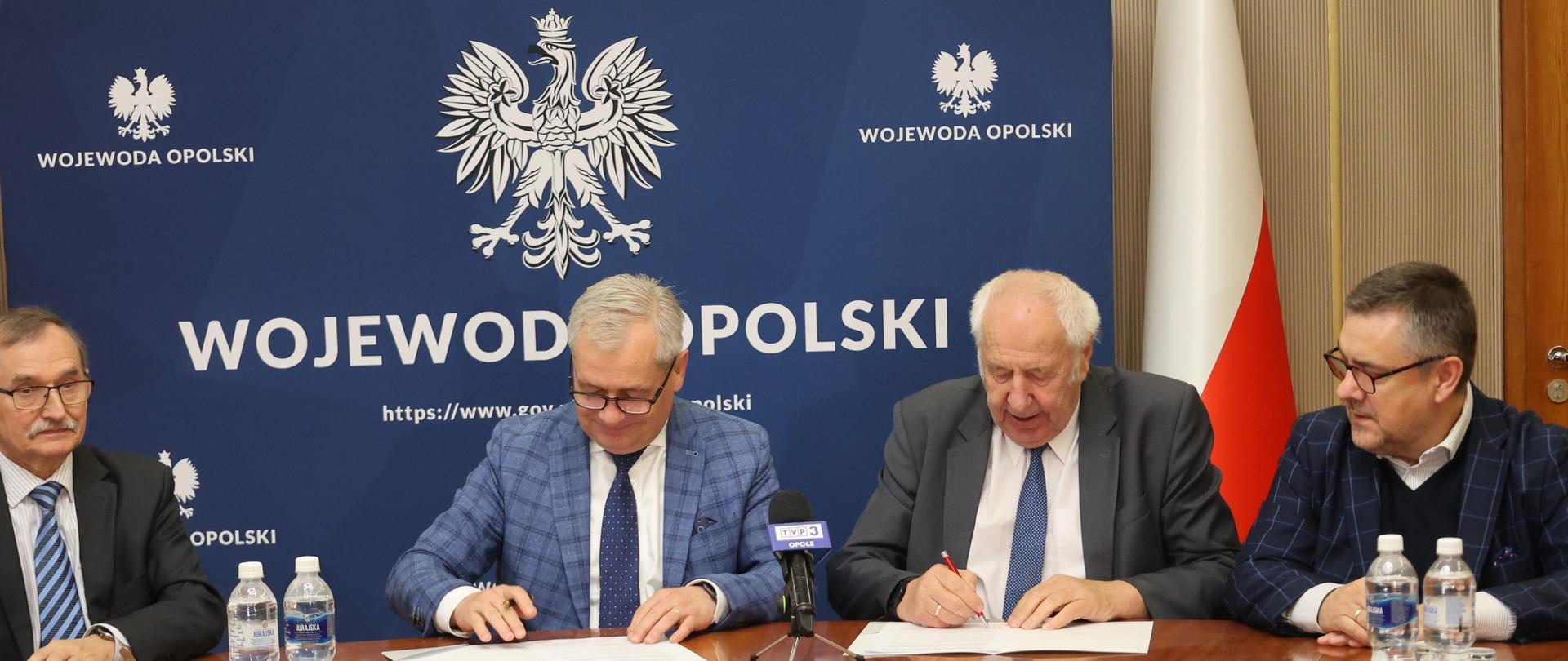 Przy stole w Sali im. Braci kowalczyków zasiadają (od lewej): Dyrektor Mieczysław Wojtaszek (Wydział Zdrowia i Polityki Społecznej), Wojewoda Opolski Sławomir Kłosowski, Starosta Powiatu Strzeleckiego Józef Swaczyna i Wicestarosta Strzelecki Waldemar Gajda. Wojewoda Opolski I Starosta Strzelecki podpisują umowę o dofinansowanie. W tle niebieska ścianka z godłem państwowym i napisem Wojewoda Opolski oraz biało-czerwona flaga.