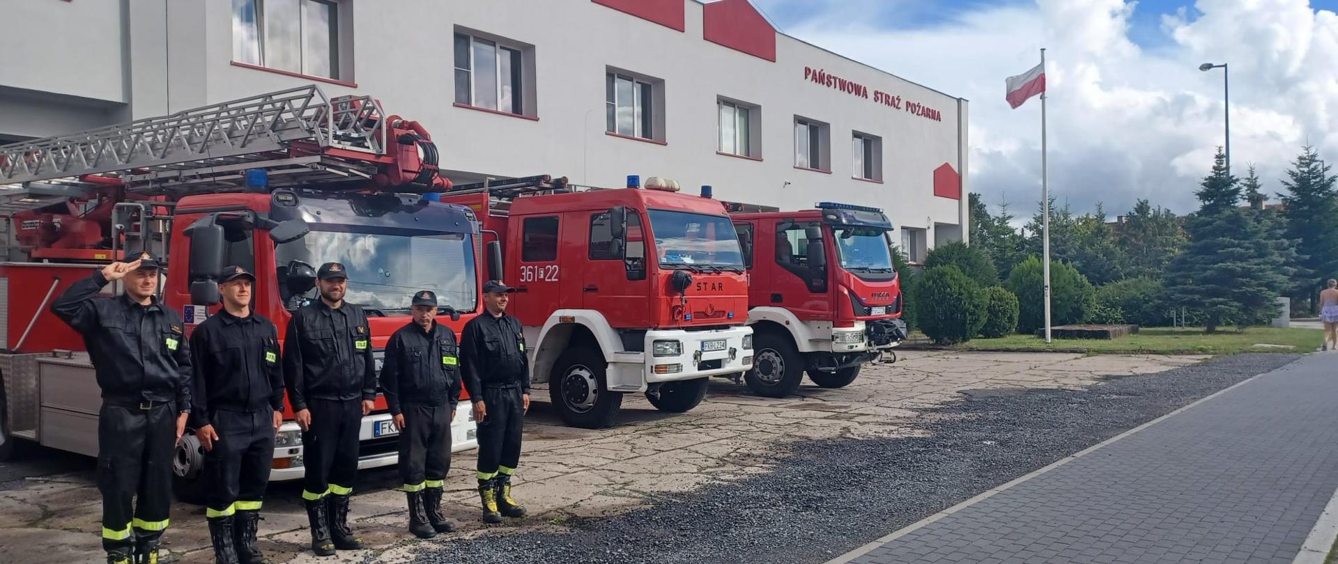 Zdjęcie przedstawia funkcjonariuszy Jednostki Ratowniczo-Gaśniczej Państwowej Straży Pożarnej w Krośnie Odrzańskim na tle pojazdów pożarniczych, którzy oddają honory Powstańcom. 