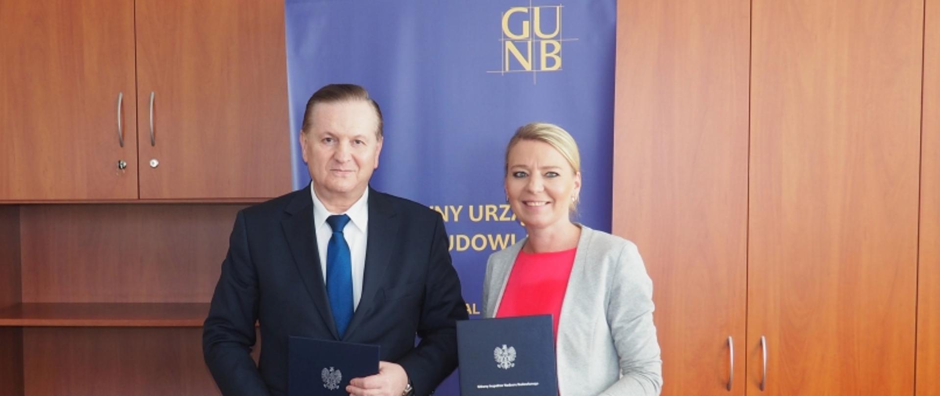Dwie osoby z teczkami z godłem stojące przed niebieskim banerem GUNB, w tle drewniane szafki.