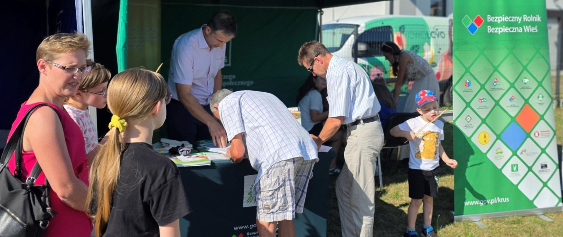 Kobieta z dziećmi i mężczyźni zgromadzeni przed namiotem, stoiskiem informacyjnym KRUS na festynie w Jabłonnie Lackiej.