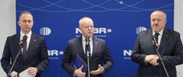 Na tle granatowej ścianki z napisem NCBR stoi przy trzech mikrofonach na stojakach trzech mężczyzn w garniturach, pośrodku wiceminister Gzik mówi do mikrofonu.