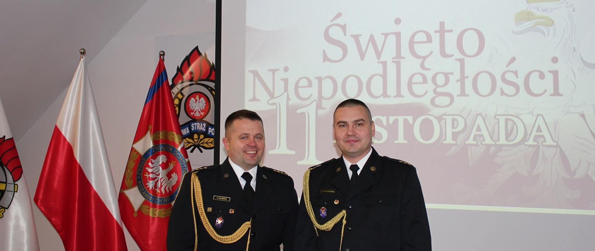 Na zdjęciu dwóch strażaków w mundurach galowych. Z lewej strony flagi. W tle napis Święto niepodległości 11 listopada.