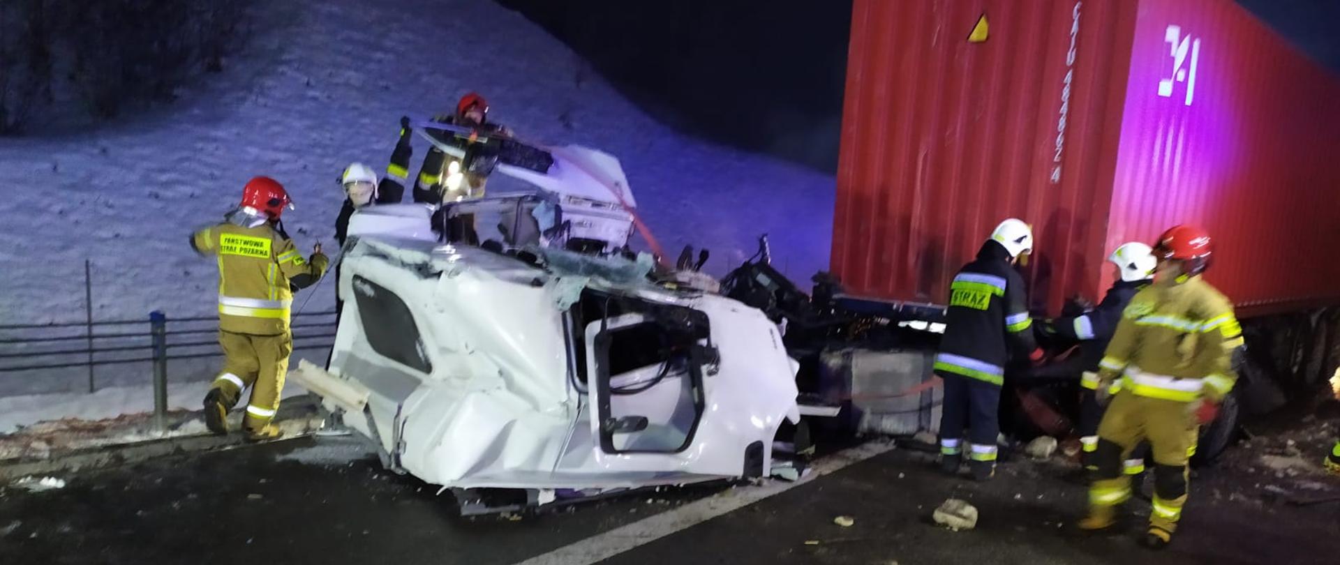 Zniszczona biała kabina pojazdu ciężarowego, leżąca przy ramie ciągnika siodłowego. Całość jest mocno odkształcona. Przy kabinie pracuje 6 strażaków w ubraniach specjalnych. W tle po lewej śnieg leżący na poboczu, po prawej czerwony, stalowy kontener