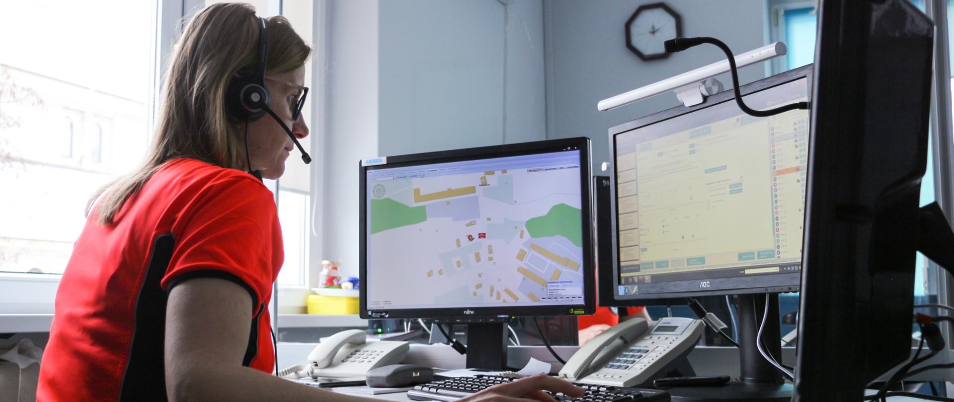 Na zdjęciu dyspozytorka medyczna, ubrana w pomarańczową bluzkę. Siedzi bokiem do obiektywu. Pracuje przy komputerze, Przed nią trzy monitory komputerowe, telefon oraz klawiatura. Kobieta na uszach ma słuchawki z mikrofonem, prowadzi rozmowę z osobą zgłaszającą zdarzenie. Kobieta ma jasne włosy oraz okulary. W tle widać okno oraz wewnętrzny parapet, na którym ulokowano środki czystości. Za monitorami komputera widać na ścianie zegar oraz klimatyzację.
