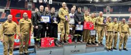 Strażacy stoją w hali sportowej. Część strażaków ma umundurowanie w kolorze piaskowym. Kilkoro strażaków ma umundurowanie w kolorze czarnym. Na środku stoją funkcjonariusze na stopniach. Na stopniach są napisane numery od 1 do 3. Na stopniu pod numerem 1 jest napis Toruń. Strażacy stojący na stopniach trzymają nagrody. W tle widać trybuny. Podłoga jest koloru niebieskiego.