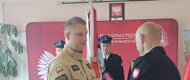 Zdjęcie przedstawia Komendanta Powiatowego PSP wręczającego strażakowi akt mianowania na wyższe stanowisko służbowe na tle sztandaru Komendy Powiatowej PSP