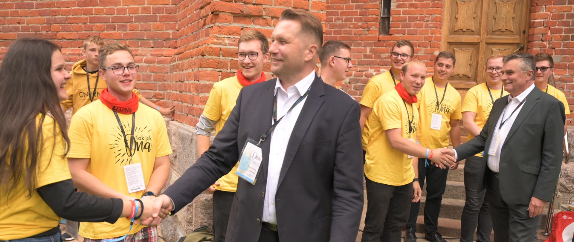 W centralnej części zdjęcia Minister Przemysław Czarnek wita się z młodzieżą zgromadzoną na spotkaniu młodych archidiecezji lubelskiej. w tle inni uczestnicy spotkania. tłem zdjęcia jest kościół.