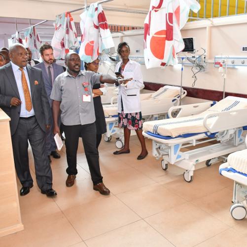 Grupa osób odwiedza oddział szpitalny, widać łóżka, jeden mężczyzna pokazuje i wyjaśnia coś grupie osób idącej za nim