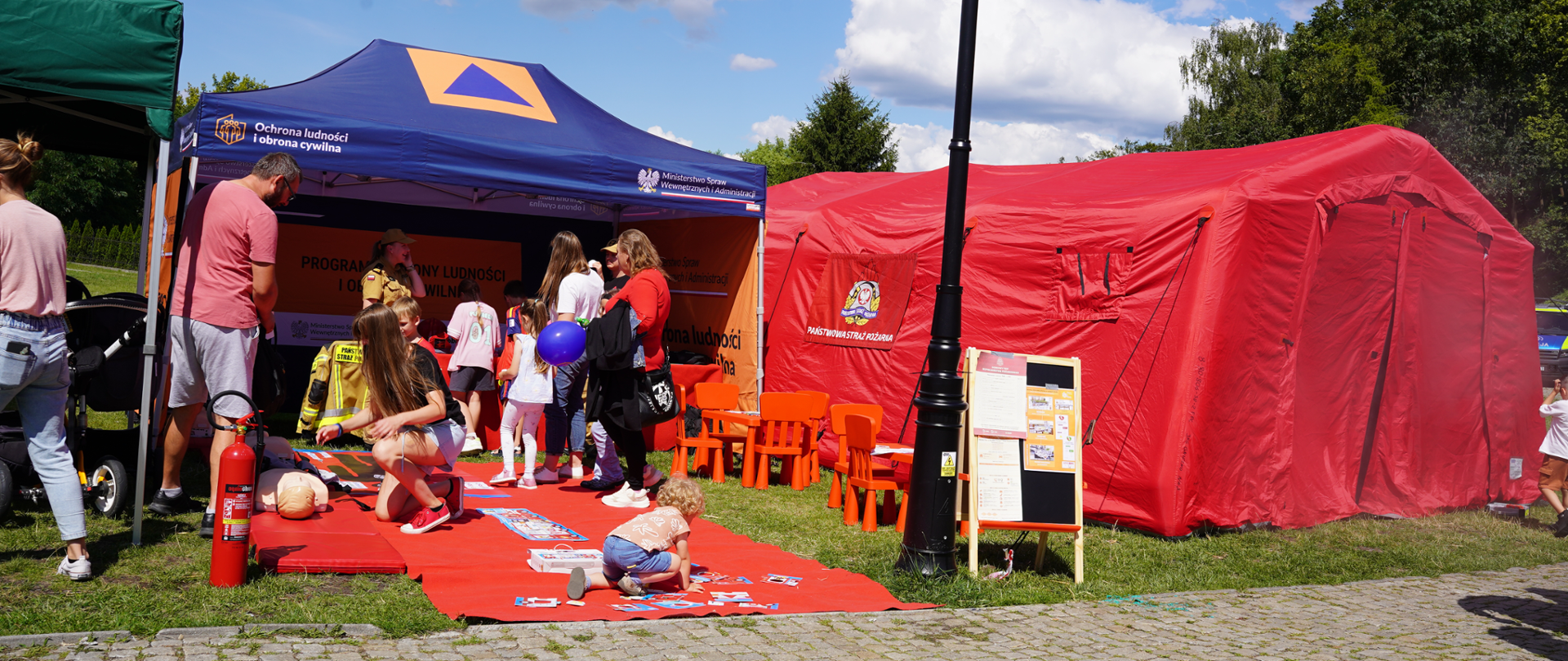 Stanowisko edukacyjne Państwowej Straży Pożarnej podczas plenerowego wydarzenia. Przed czerwonym namiotem PSP i niebieskim namiotem programu ochrony ludności dzieci biorą udział w zabawach i grach edukacyjnych, a dorośli odwiedzają stoisko. Na pierwszym planie widoczne są czerwona mata do zajęć, gaśnica oraz tablica informacyjna