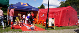 Stanowisko edukacyjne Państwowej Straży Pożarnej podczas plenerowego wydarzenia. Przed czerwonym namiotem PSP i niebieskim namiotem programu ochrony ludności dzieci biorą udział w zabawach i grach edukacyjnych, a dorośli odwiedzają stoisko. Na pierwszym planie widoczne są czerwona mata do zajęć, gaśnica oraz tablica informacyjna