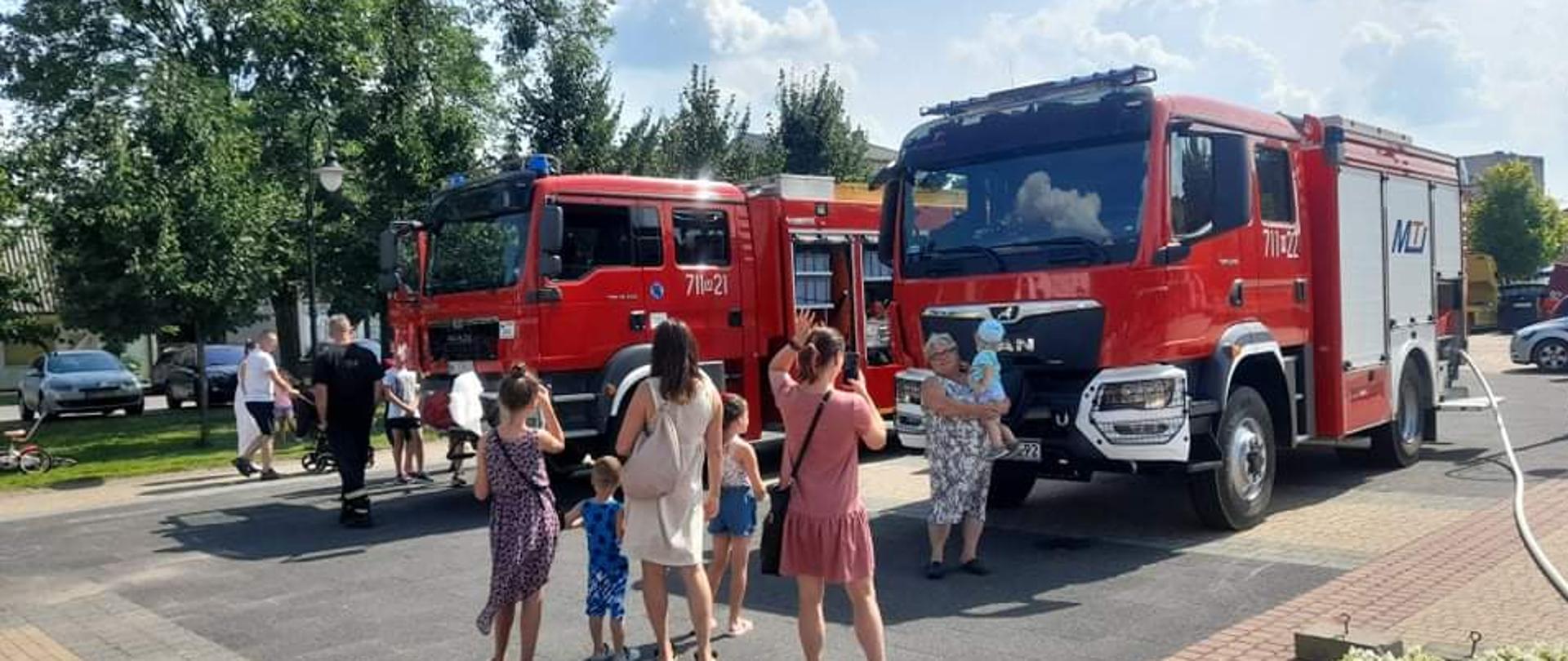 Dwa samochodu ratowniczo- gaśnicze stoją w środku parku. Wokół nich stoją ludzie z dziećmi oglądający sprzęt strażacki.