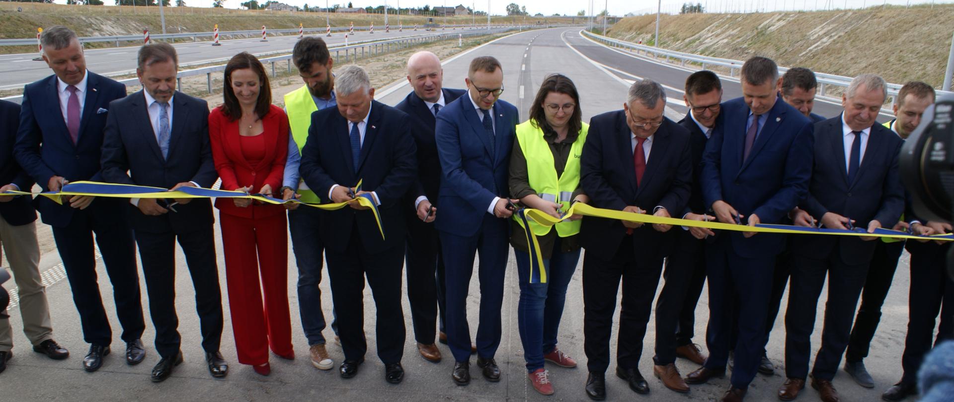 uczestnicy uroczystości stoją przed przecinaną wstęgą, wstęga została przed chwilą przecięta w jednym miejscu