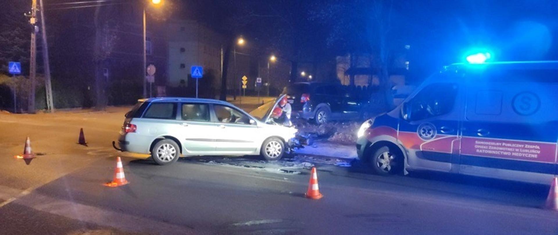 Zdjęcie zrobione na tle zabudowań w Lublińcu na skrzyżowaniu ulic Żwirki i Wigury oraz Sobieskiego. W centralnej części widać bok samochodu osobowego po wypadku z podniesioną maską komory silnika przy której pracuje dwóch strażaków w ubraniach specjalnych w hełmach. Po prawej stronie stoi samochód Zespołu Ratownictwa Medycznego a w oddali, na chodniku stoi drugi samochód osobowy biorący udział w kolizji.