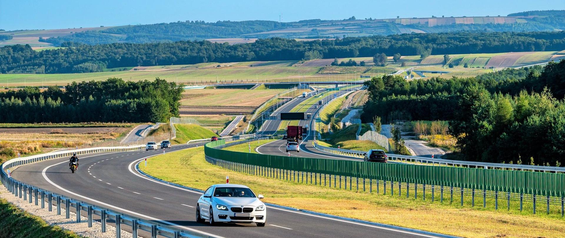 Droga ekspresowa S7 na odcinku Moczydło - Miechów. Dwujezdniowa droga ekspresowa wijąca się między polami i lasami po niewielkich pagórkach. Widoczne pojazdy poruszające się w obu kierunkach.