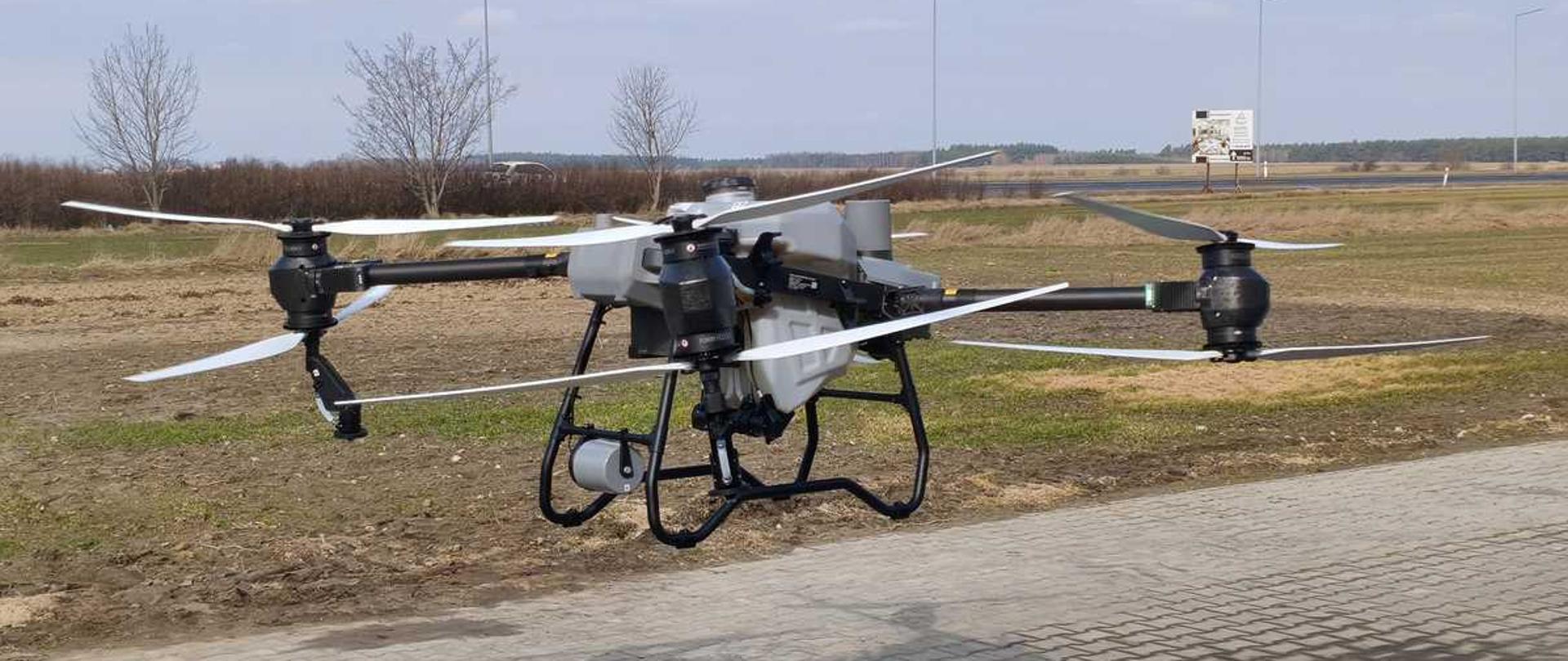 Poziome zdjęcie przedstawiające duży, profesjonalny dron rolniczy (quadrocopter), który unosi się nisko nad ziemią, tuż przy krawędzi wybrukowanego kostką placu.Dron ma masywny, szary korpus z centralnie umieszczonym, dużym zbiornikiem na płyny (np. do oprysków). Od korpusu odchodzą cztery długie, czarne ramiona z potężnymi silnikami i dużymi, białymi śmigłami, które są w ruchu. Cała konstrukcja opiera się od spodu na czarnym, metalowym podwoziu w formie płozy.W tle rozciąga się otwarty, płaski krajobraz polno-łąkowy z kępami suchych, bezlistnych drzew i krzewów. W oddali, po prawej stronie, widoczny jest pionowy baner reklamowy przy drodze oraz rząd wysokich latarni ulicznych ciągnących się wzdłuż horyzontu. Nad całością roztacza się jasnoniebieskie niebo z delikatnymi, białymi chmurami.
