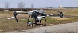 Poziome zdjęcie przedstawiające duży, profesjonalny dron rolniczy (quadrocopter), który unosi się nisko nad ziemią, tuż przy krawędzi wybrukowanego kostką placu.Dron ma masywny, szary korpus z centralnie umieszczonym, dużym zbiornikiem na płyny (np. do oprysków). Od korpusu odchodzą cztery długie, czarne ramiona z potężnymi silnikami i dużymi, białymi śmigłami, które są w ruchu. Cała konstrukcja opiera się od spodu na czarnym, metalowym podwoziu w formie płozy.W tle rozciąga się otwarty, płaski krajobraz polno-łąkowy z kępami suchych, bezlistnych drzew i krzewów. W oddali, po prawej stronie, widoczny jest pionowy baner reklamowy przy drodze oraz rząd wysokich latarni ulicznych ciągnących się wzdłuż horyzontu. Nad całością roztacza się jasnoniebieskie niebo z delikatnymi, białymi chmurami.