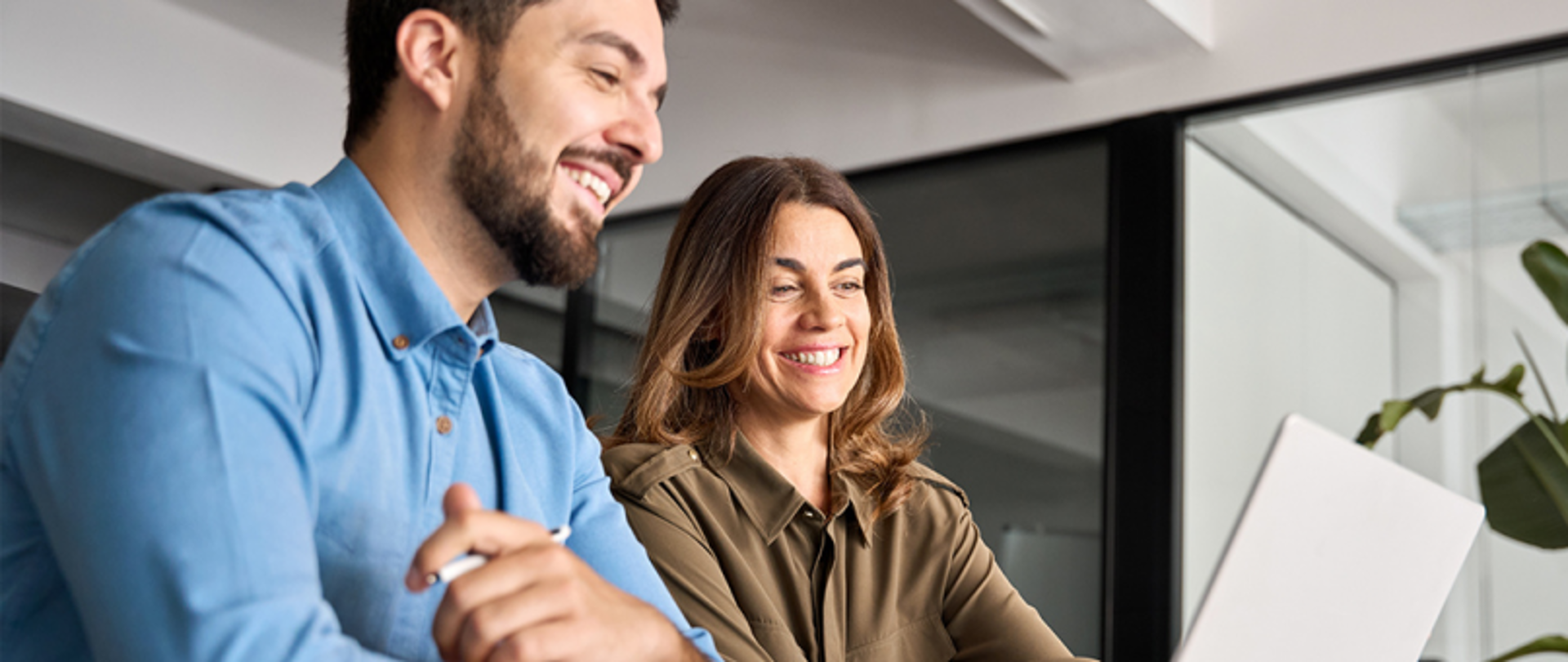 Na zdjęciu dwie uśmiechnięte osoby, prowadzą szkolenie – kobieta i mężczyzna – siedzącą przy biurku przed ekranem laptopa.