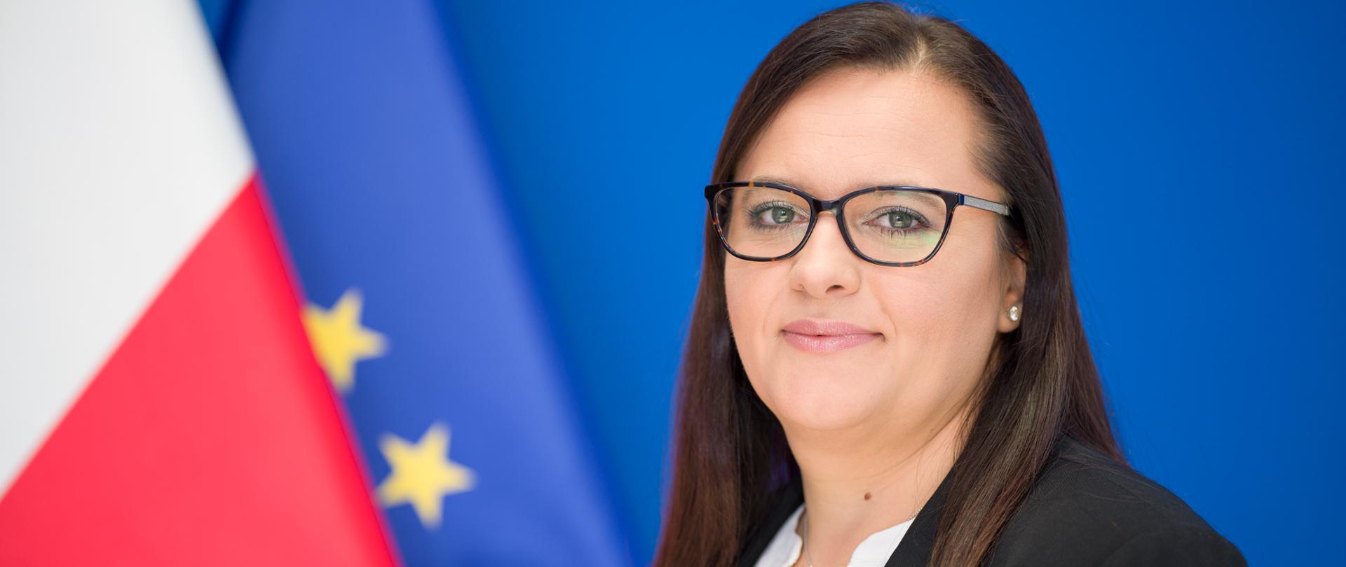 minister Małgorzata Jarosińska-Jedynak patrzy w stronę fotografującego, za nią flagi Polski i Unii Europejskiej