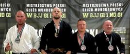 Czterech mężczyzn w strojach do BJJ stoi na podium podczas dekoracji zwycięzców Mistrzostw Polski Służb Mundurowych. Zawodnik na najwyższym stopniu podium trzyma koszulkę z napisem "Państwowa Straż Pożarna". Wszyscy mają na szyjach medale, w tle baner zawodów.