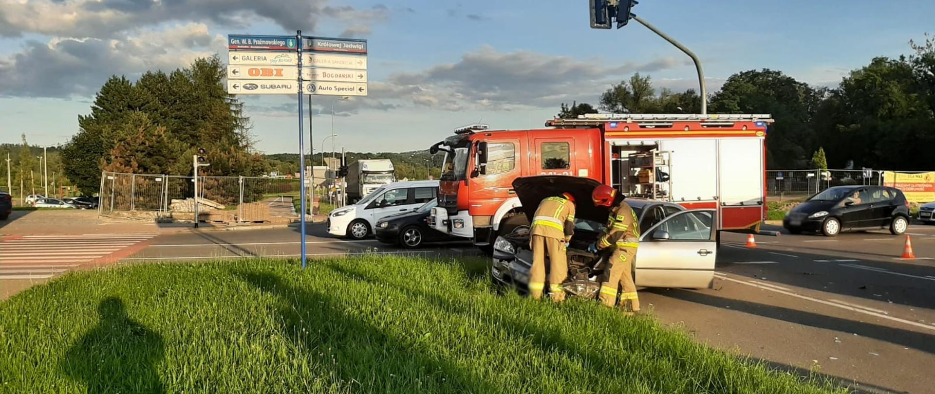 Strażacy wykonują czynności pod otwartą maską silnika samochodu osobowego. W tle samochód pożarniczy.