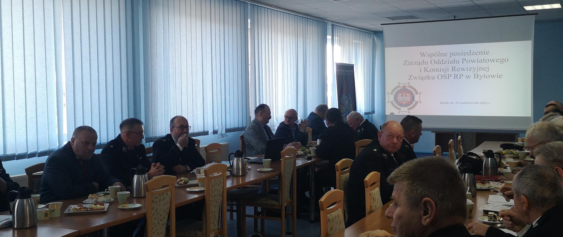 Zdjęcie przedstawia mężczyzn w umundurowaniu galowym siedzących przy okrągłym stole i oglądających wyświetloną prezentację multimedialną. 