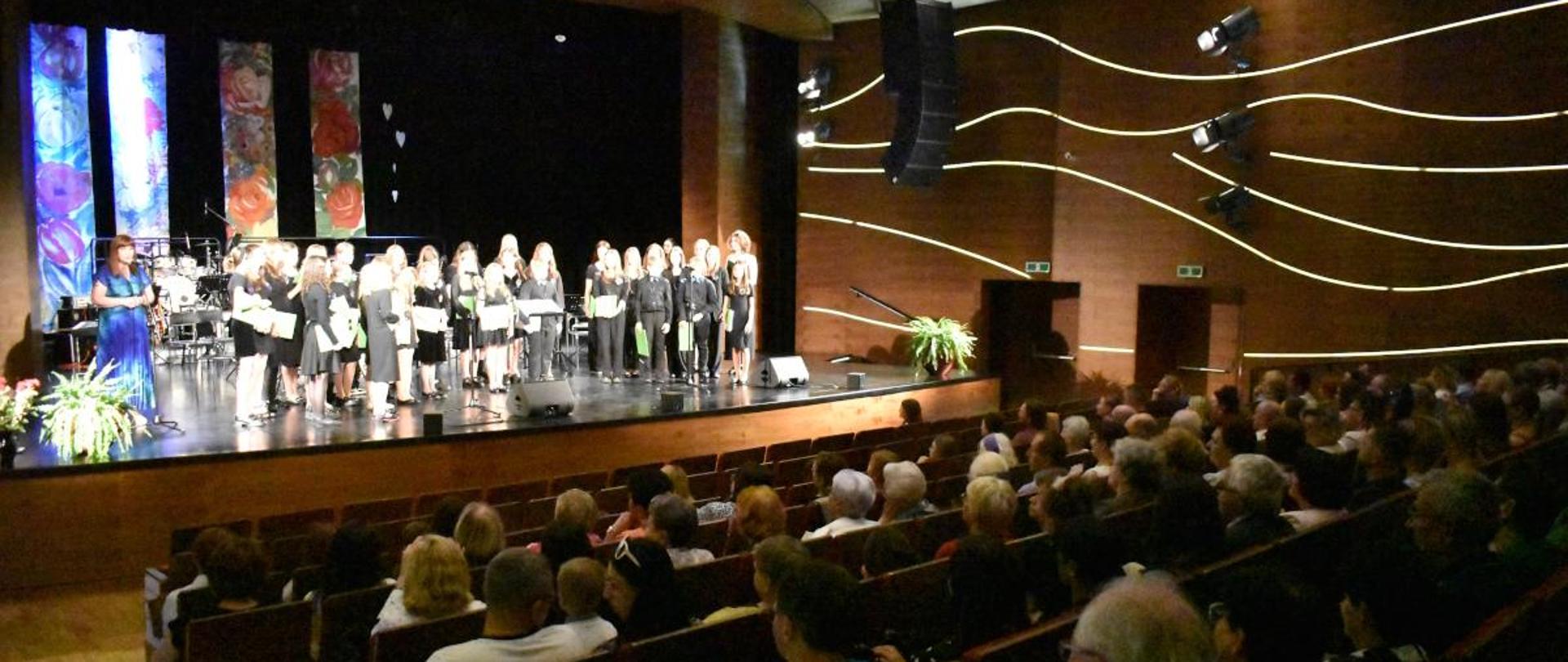 na zdjęciu, w sali widowiskowej JDK, podczas Koncertu dla Mam, na drugim planie widnieje na scenie chór szkolny i dyrektor PSM I st. w Jaśle. Na pierwszym planie widnieje publiczność, która zasiada na widowni. Kolorystyka zdjęcia jest biało-brązowo- szaro-niebiesko-czarna.