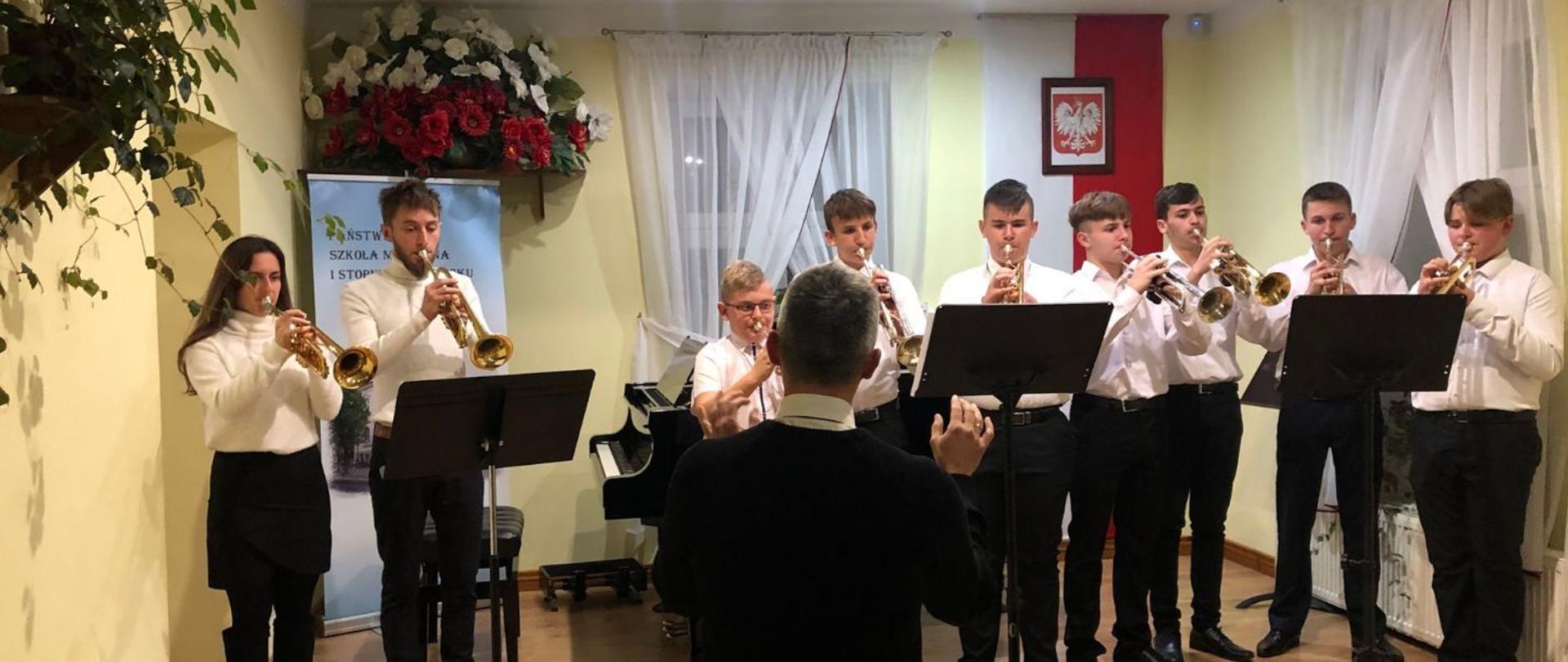 Zespół trębaczy na scenie. Za zespołem po lewej kwiatowa aranżacja w kolorach biało-czerwonych, poniżej banner szkoły muzycznej. W centrum za zespołem fortepian a po prawej pionowo rozwieszona flaga narodowa, na niej obraz z godłem Polski.