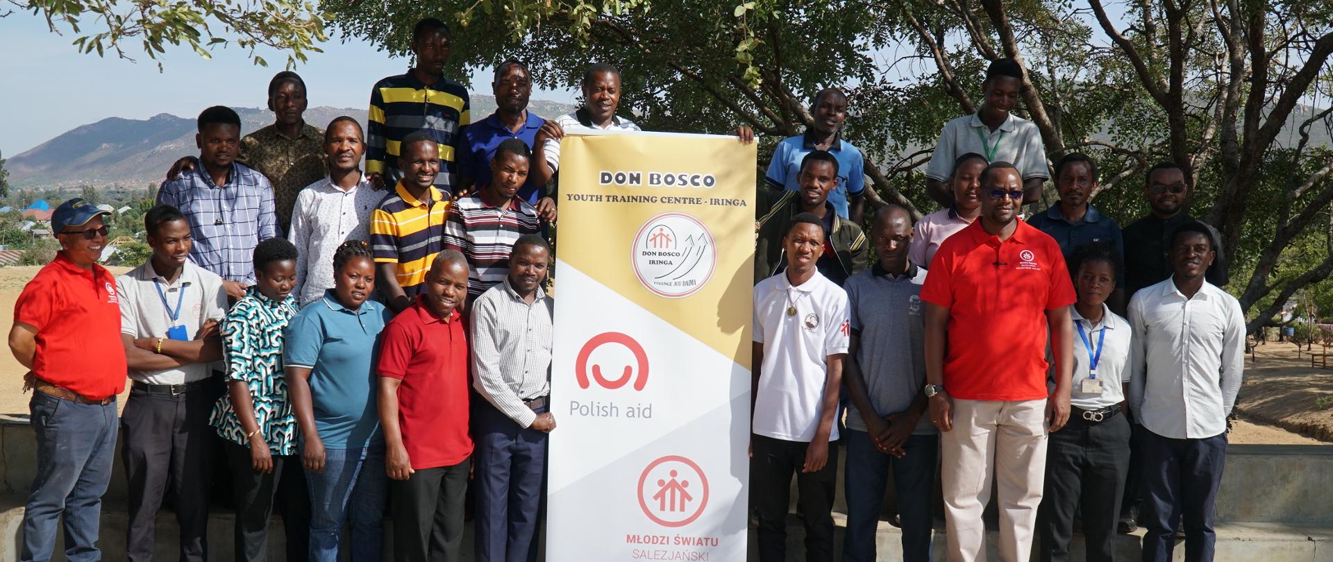 Pod drzewem stoją w trzech rzędach nauczyciele i nauczycielki, z których część trzyma baner informacyjny szkoły