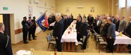 Zdjęcie przedstawia strażaków i osoby zaproszone, które znajdują się wewnątrz pomieszczenia (na sali). Osoby znajdują się w pozycji stojącej zwrócone w kierunku sztandaru.