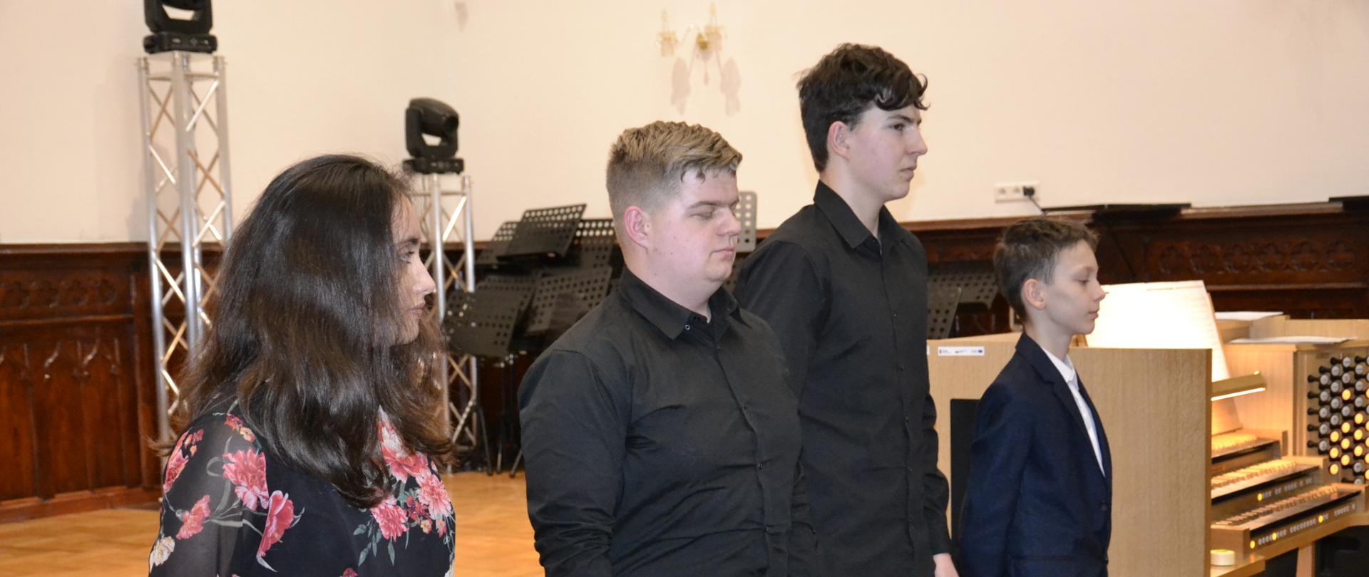 zdjęcie przedstawia czterech uczniów klasy organów naszej szkoły, a za nimi blado-kremowe organy oraz sceną Sali Królewskiej Państwowej Szkoły Muzycznej w MIelcu