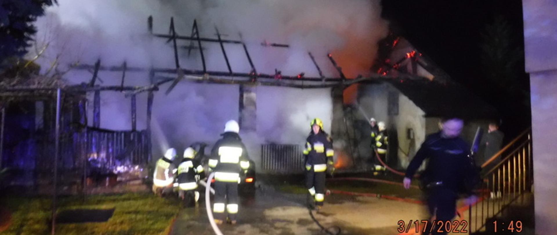 Zdjęcie przedstawia budynek gospodarczy w którym pali się dach. Jest pora nocna. Strażacy podają dwa prądy gaśnicze wody na obiekt. Przez teren prowadzonej akcji poprowadzone są linie wężowe, które służyły do podania środka gaśniczego na palące się obiekty. Strażacy znajdują się na terenie przed płonącymi obiektami , prowadząc działania gaśnicze.
