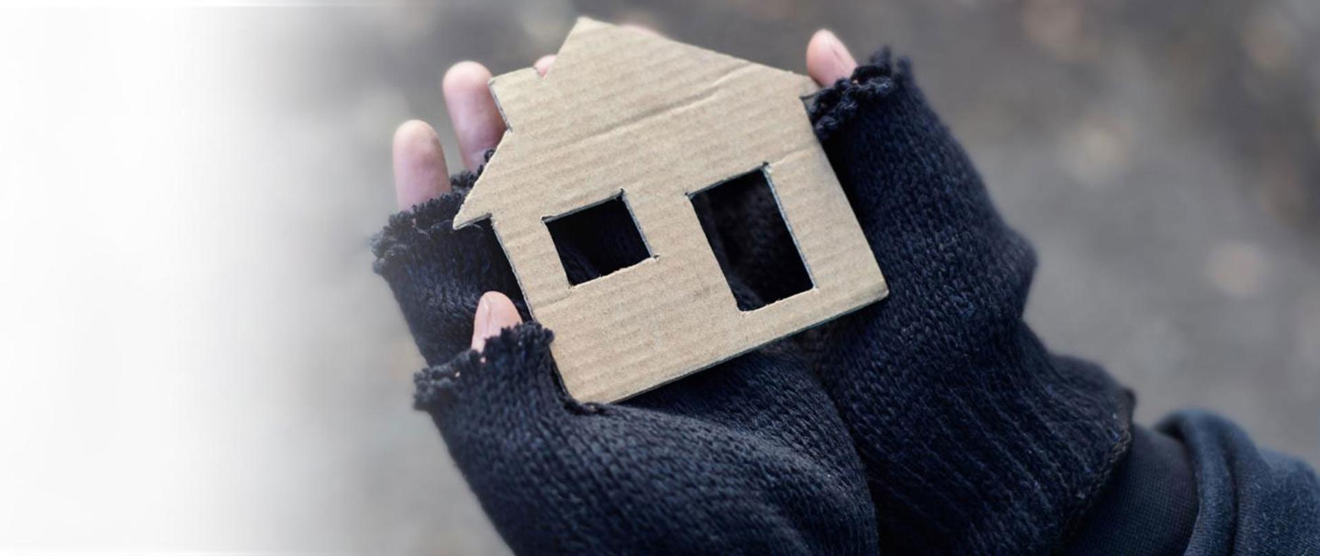 Na zdjęciu dłonie w wełnianych rękawiczkach, trzymające wycięty z kartonu kształt domu.