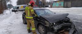 Na pierwszym planie widać strażaka stojącego przy uszkodzonym samochodzie osobowym. Natomiast na drugim planie widać drugi uszkodzony samochód, biorący udział w kolizji oraz samochód ratowniczo gaśniczy.