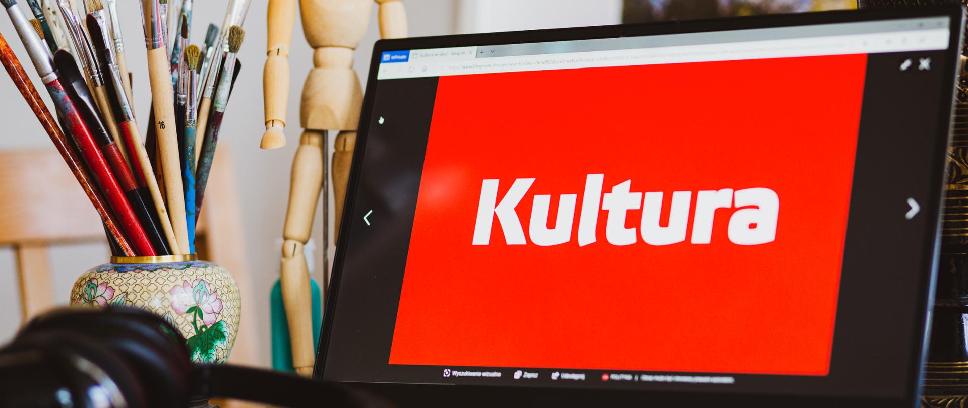 Laptop z białym napisem kultura na czerwonym tle wyświetlanym na ekranie, w tle mały dzbanek z pędzlami i drewniana kukiełka, fot. Danuta Matloch 