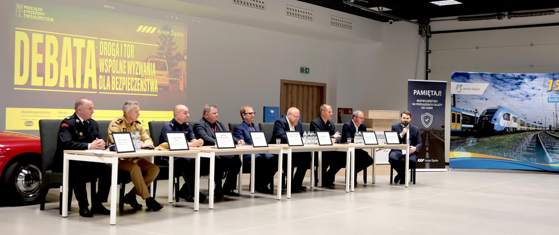 To zdjęcie przedstawia oficjalną debatę lub panel dyskusyjny odbywający się w dużej, nowoczesnej sali konferencyjnej. Przy długim rzędzie ustawionych obok siebie stołów siedzi dziewięciu mężczyzn. Większość z nich ma na sobie garnitury, ale kilku występuje w mundurach służb. Jeden z uczestników, siedzący po prawej stronie, trzyma mikrofon i zabiera głos. Przed każdym uczestnikiem znajduje się tabliczka z imieniem i nazwiskiem. W tle, na dużym ekranie projekcyjnym, wyświetlony jest slajd w kolorach żółtym i czarnym. Widnieje na nim duży napis „DEBATA” oraz hasło dotyczące wspólnych wyzwań dla bezpieczeństwa na drogach i torach kolejowych. Widać również logo przewoźnika kolejowego oraz elementy związane z kampanią bezpieczeństwa na przejazdach kolejowych. Po prawej stronie sali stoją dwa duże banery. Jeden przypomina o bezpieczeństwie na przejazdach kolejowych, drugi przedstawia nowoczesny pociąg na torach pod błękitnym niebem. Na lewym skraju kadru widać fragment czerwonego samochodu, prawdopodobnie element ekspozycji. Sala jest jasna i przestronna, z jasną podłogą oraz wysokim sufitem. 