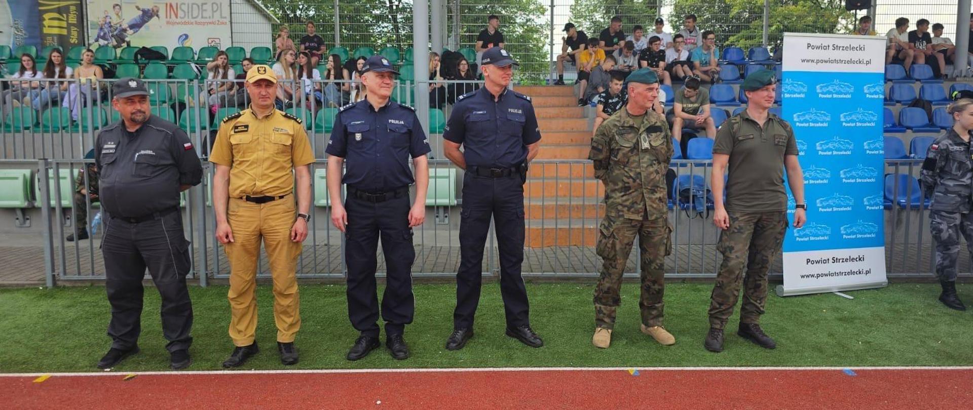 Na zdjęciu znajduje się strażak, policjanci, żołnierze, funkcjonariusze SW w umundurowaniu służbowym. W tle widać trybuny stadionu i kibice