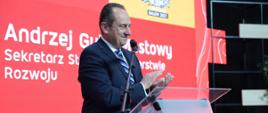 Wiceminister rozwoju i technologii Andrzej Gut-Mostowy podczas otwarcia TOUR SALON w Poznaniu, wiceminister stoi za mównicą i klaszcze w dłonie