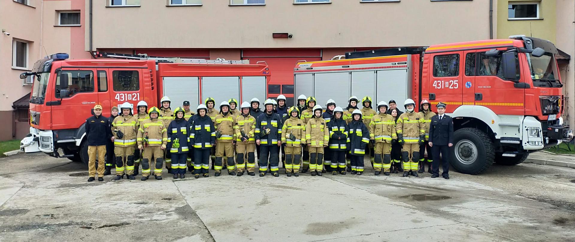 Na dworze na tle dwóch wozów bojowych stoi kilkudziesięciu strażaków w dwuszeregu w ubraniach bojowych.