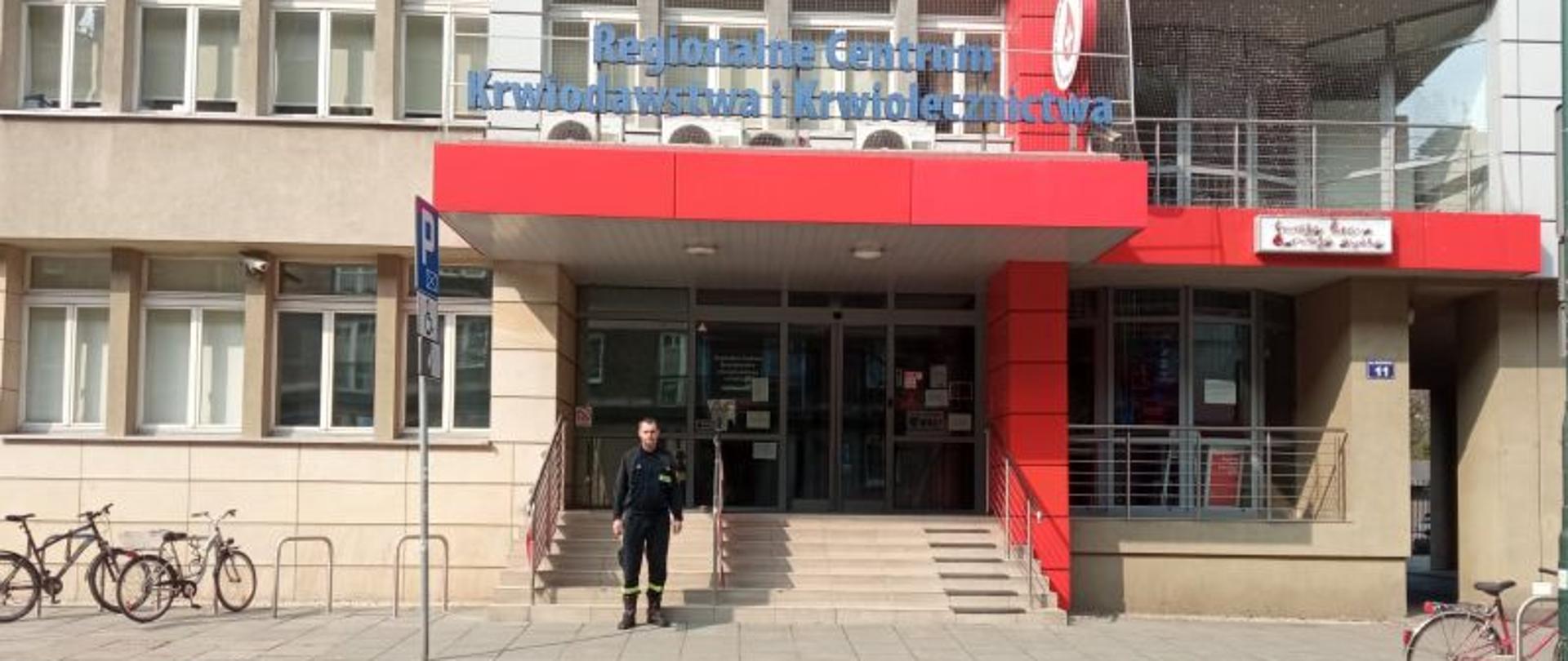 Zdjęcie przedstawia ogniomistrza Tadeusza Szopińskiego w umundurowaniu koszarowym stojącego przed wejściem głównym do Regionalnego Centrum Krwiodawstwa i Krwiolecznictwa w Krakowie